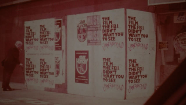 Pink toned still of a man approaching a wall papered over with posters reading "The film the FBI doesn't want you to see"