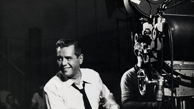 A smiling Desi Arnaz, sitting next to a film camera, looks out of frame
