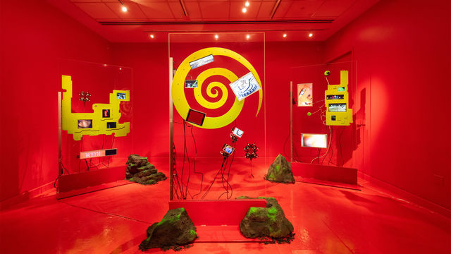 Installation image of a red room with sculptures made of transparent glass, yellow shapes, and electronic components