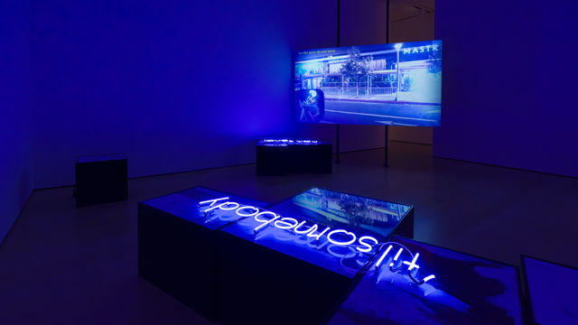 Installation with a video screen in the background showing a street scene, and a neon sign reading "til' somebody" laying on a plinth in the foreground