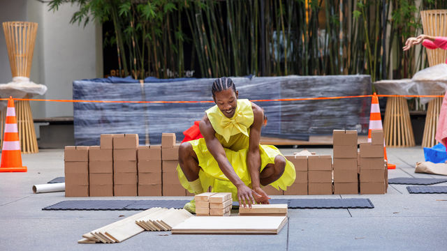 A dancer in a yellow outfit smiles as he kneels in front a stack of wood pieces