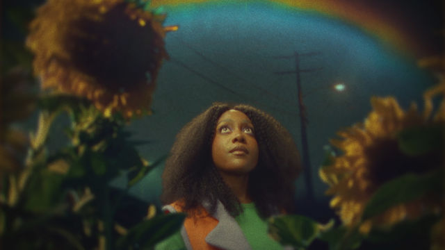 A Black woman, framed by sunflowers, looks up at a rainbow in the night sky