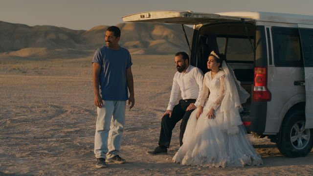 A couple dressed for their wedding sits in the open hatch of a van in the dessert