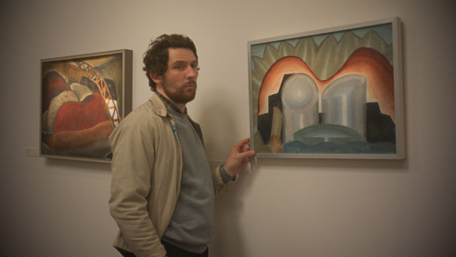 A man in a tan jacket fingers a framed painting