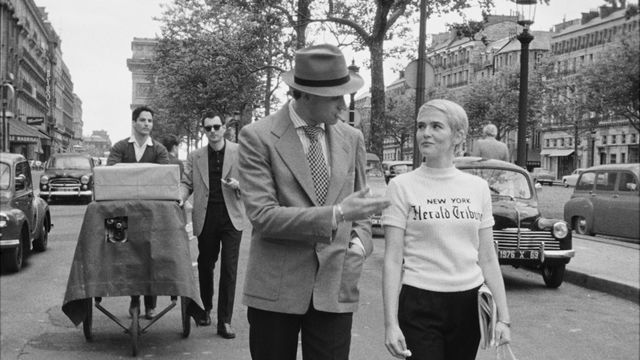 A man and a suit and a woman in a New York Herald Tribune shirt walk down a city street, followed by a cameraman