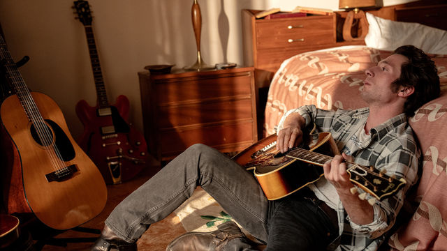 Jeffrey Allen White as Bruce Springsteen sits against a bed playing an acoustic guitar