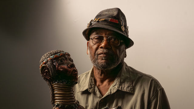 The artists Akinsanya Kambon sits next to his sculpture of a long-necked figure