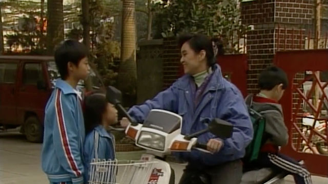 A woman on a motor scooter talks to two children in matching tracksuits