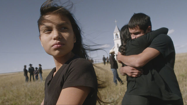 A girl looks off-camera, while behind her a man and woman hug in front of a church