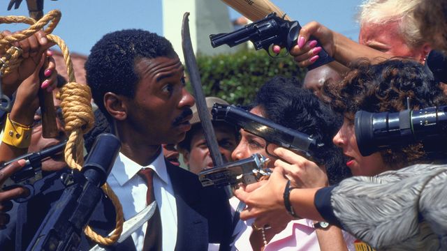 A Black man in a suit is surrounded by people pointing weapons at him