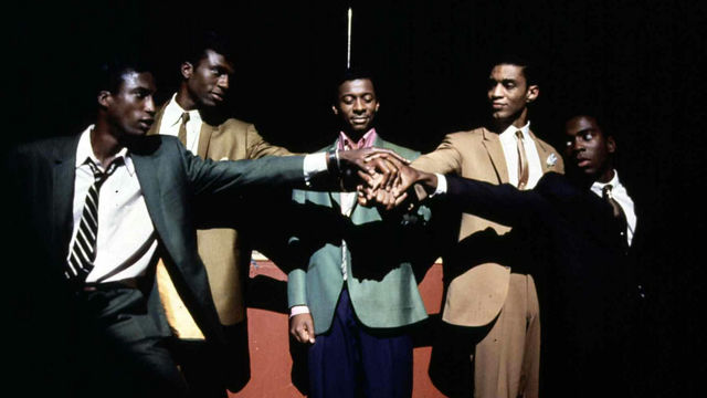 Five Black men stand in a semi-circle, their hands joined in the center