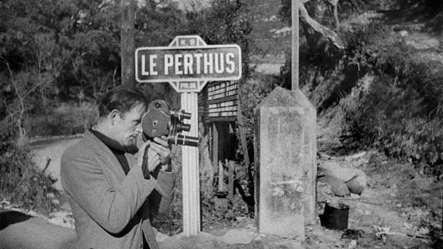 A man loks through a small, handheld film camera as he stands in front of a sign reading "LE PERTHUS"