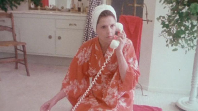 A woman in a pink robe kneels on the phone as she talks into a telephone