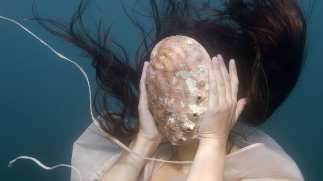 A woman, underwater, holds a large shell in front of her face