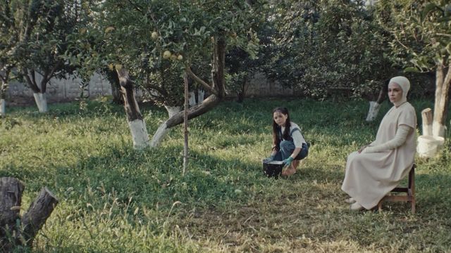 A girl kneels next to a woman in all white amidst a garden