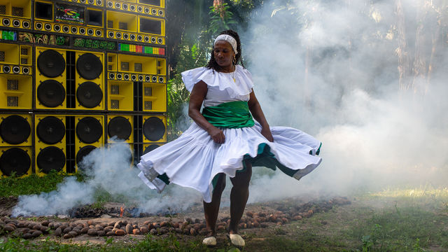 A woman in a white dress dances in front of a wall of yellow speakers, surrounded by smoke