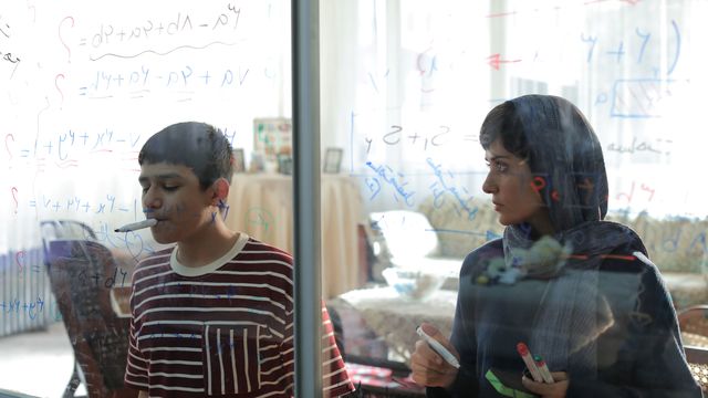 A woman and a boy stand in front of a clear writing board covered in math equations, where the boy draws with a marker in his mouth