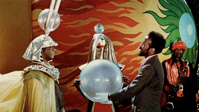 Sun Ra, in an elaborate headdress, stands with a man holding a large orb in his white-gloved hands