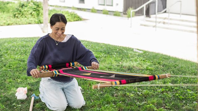 A woman kneels on the ground, holding a weaving suspended in front of her