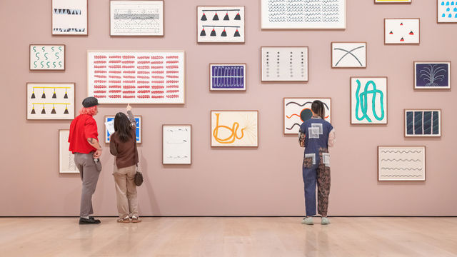 Three visitors, viewed from behind, stand in front of a pink gallery wall covered in a cluster of framed abstract artworks