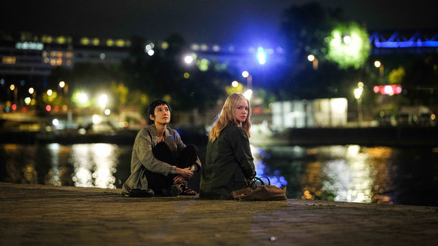 Two women sit on the waterfront, with city lights twinkling behind them