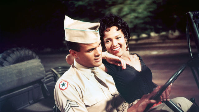 A woman in a black dress and a man in a solider's uniform sit in the front seat of a jeep, the man holding the steering wheel while the woman rests her elbow on his shoulder and smiles