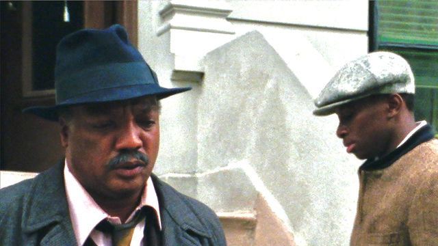Two men, one facing the camera wearing a fedora and suit, the other facing down