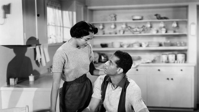 A woman stands over a man sitting at a table, pencil in hand, in a kitchen