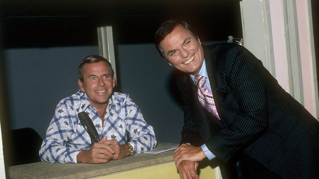 A man in a pin-striped suit smiles as he leans against a desk, where another smiling man sits in front of a microphone