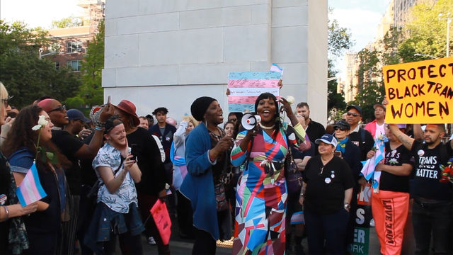 A dark skinned person with long black hair and a colorful dress speaks into a loudspeaker at an outdoor protest