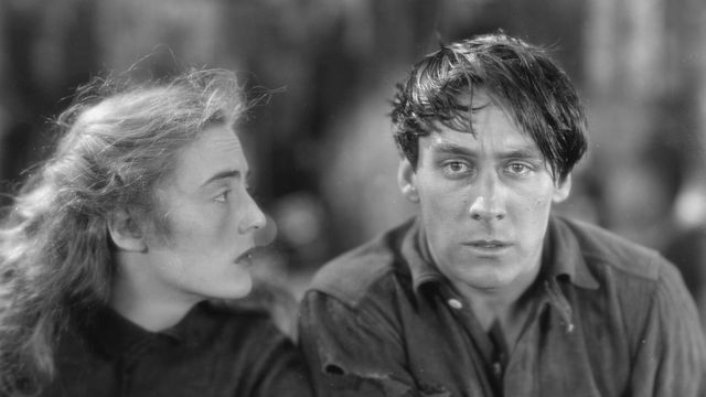 On the right, a man with dark hair swept across his forehead looks at the camera, while a woman on the left gazes at him