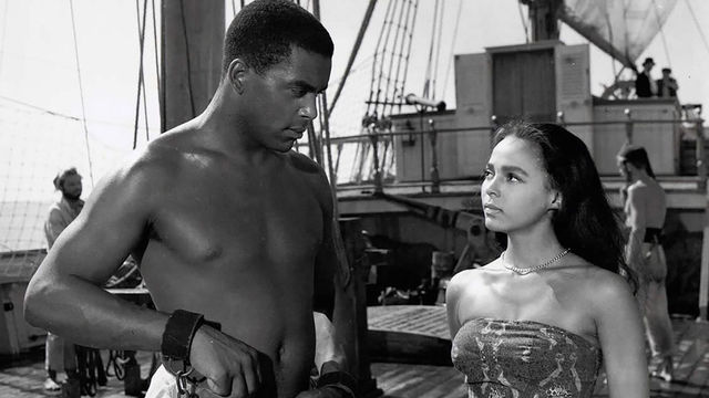 A man and woman looking at each other on a ship. The man is enslaved in shackles.