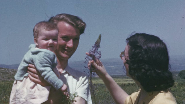 A man holds a baby in his arms, while a woman holds up a large purple flower