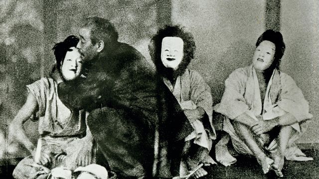 Three figures sit wearing white masks, while a man embraces the figure on the left