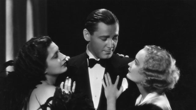 A dapper man in a tuxedo looks at a blond woman clinging to his left arm, while a dark haired woman clings to his right