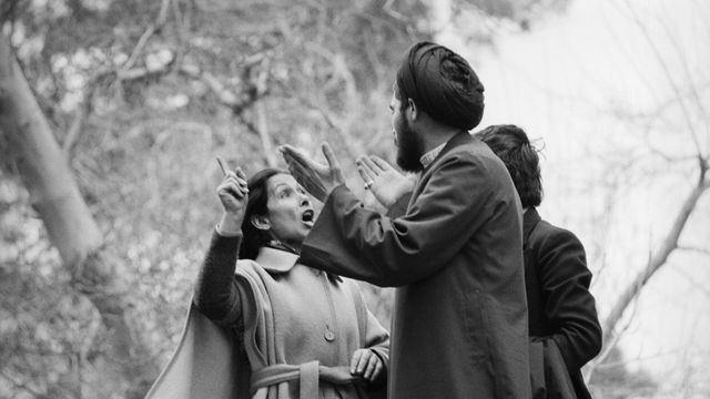 A woman in a trench coat in animated conversation with a man in a head wrap