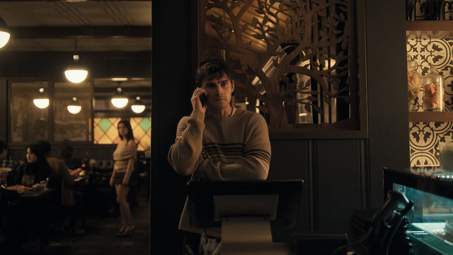 A young man in a striped sweater holds a cell phone next to his head, while out-of-focus in the background, a long-haired figure in a beige sweater and dark skirt looks his way