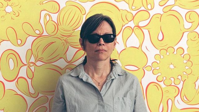 Photo of a woman wearing sunglasses and standing against a yellow flowered background 