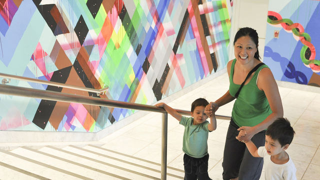 Photo of a set of white stairs leading up to the left with a railing in the center. A person with dark hair holds two small people by each hand while walking up the stairs and smiling. The walls in the background re painted with bright geometric colors in blues, greens, pinks, yellows and browns.