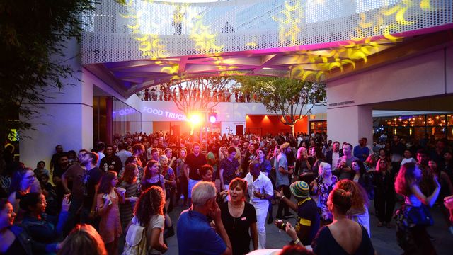 An open air courtyard at dusk, with many people filling the bottom one-third of the frame. Pink and gold lights reflect off the heads of the people. A bright, yellow light is just to the left of the middle of the image. At the top of the image, a perforated structure spans the space above the courtyard. The bottom of the structure is lit with pink from below, and yellow lights in the shape of symbols or numbers.