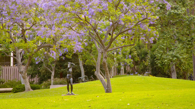 A large grassy area with trees in the middle ground blooming with purple flowers, and a single bronze sculpture of a nude woman standing on the grass in the middle ground, and another abstract sculpture in the background. More trees are in the background, and appear to obscure buildings.