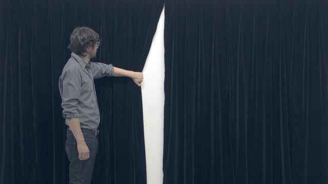 Aman stands in front of blue curtains, and is reaching out with arm to pull back one of the curtains slightly revealing white space.