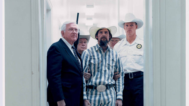 Men stand in a white doorway, all wearing white cowboy hats. The man in the center foreground wears a striped outfit and is bearded. A man in the background holds a shotgun.