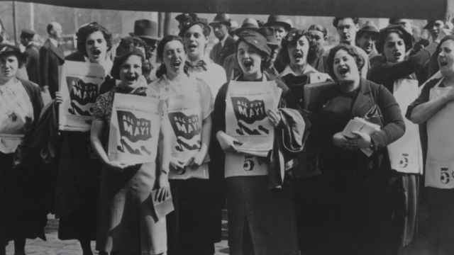 Black and white photo shows a crowd of women standing, facing the camera, with their mouths open as if shouting. Many women in the foreground hold papers against their chests that have a graphic of a flag with "All Out May 1" written on the flag.