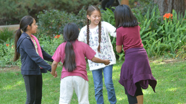 Children stand on a grassy area in a circle, holding hands and smiling, with plants in the background. 