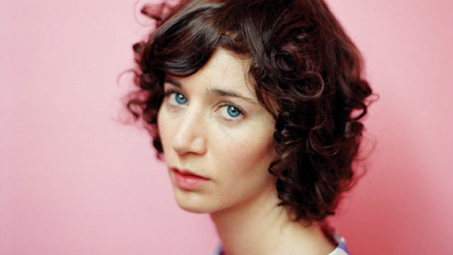Photpgraph of a light-skinned person in 3/4 view with neck-length dark hair, wearing a striped shurt on a pink background.
