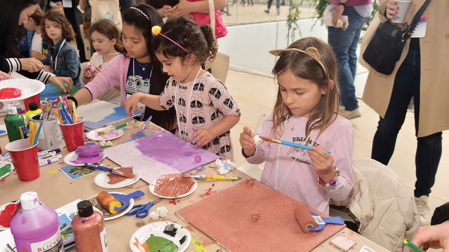 Several children stand at work at a table with art supplies, paint, and paintbrushes.