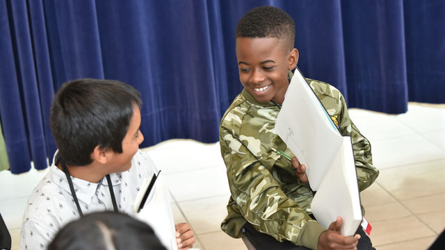 Two children holding notebooks and pencils look at each other, smiling. One holds up his notebook to show the other a drawing.