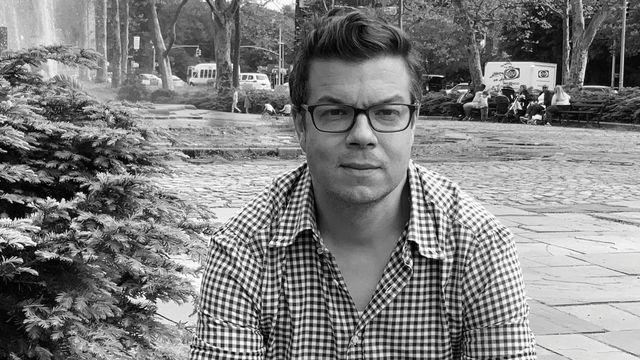 Black and white photo of Ben Lerner sitting in an outdoor city scene. In the background are trees, people sitting on benches and a street with cars.