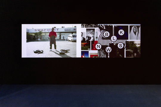 A photograph of two videos projected side-by-side against a black wall that takes up two-thirds of the image. The carpet in the lower third of the photograph is blue.